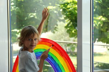 Little boy drawing rainbow on window. Stay at home concept clipart