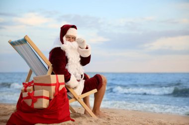 Noel Baba hediyelerle dolu bir çanta kumsalda sandalyede dinleniyor, mesaj atmak için yer var. Noel tatili