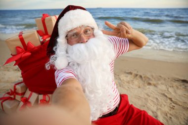 Noel Baba bir çanta dolusu hediyeyle sahilde selfie çekiyor. Noel tatili