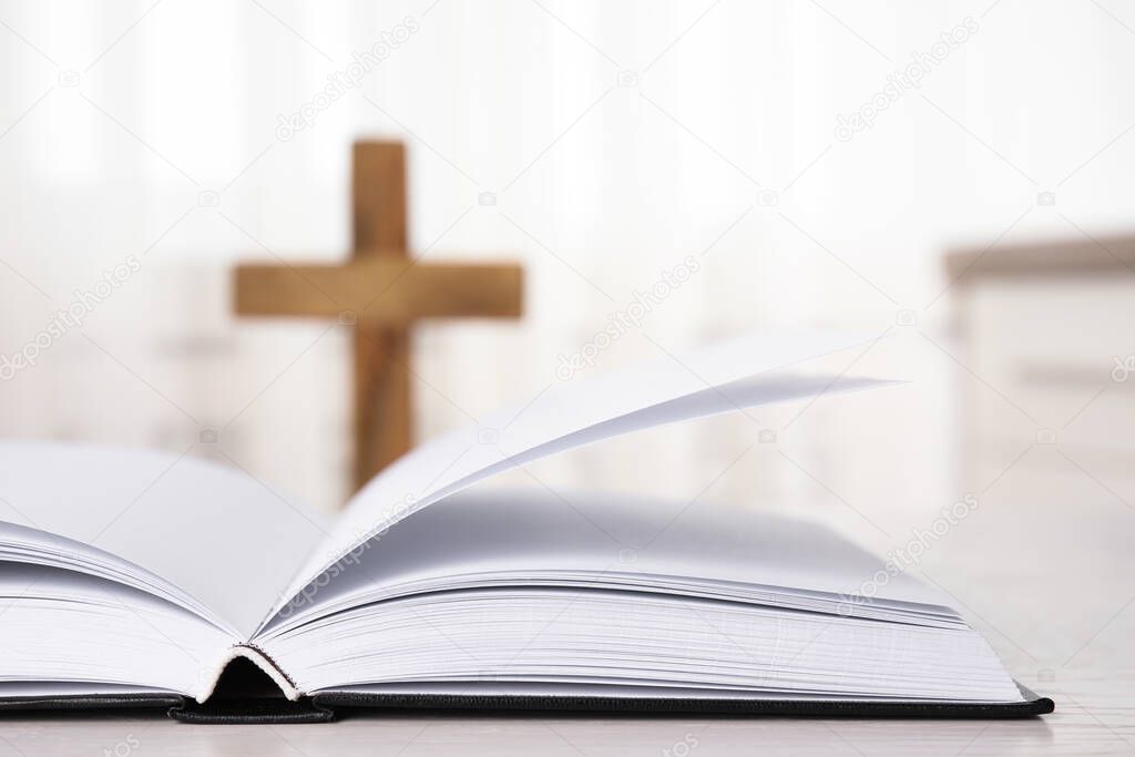 Open Bible on table and blurred cross on background, closeup. Christian religion