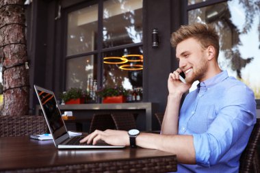 Açık kafede dizüstü bilgisayar kullanırken telefonla konuşan mutlu genç adam.