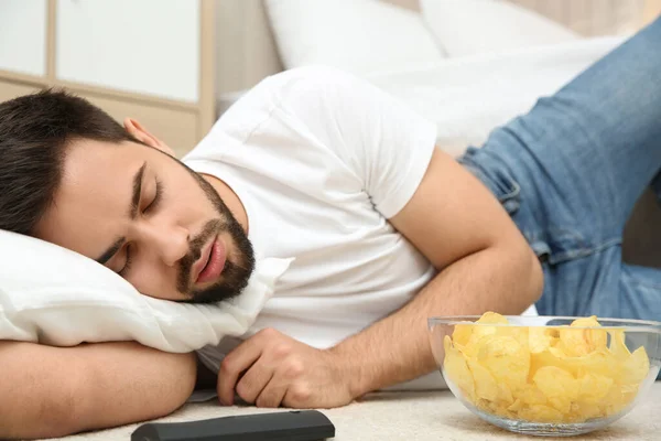 一个懒惰的年轻人 带着一碗薯片和电视遥控器睡在家里的地板上 — 图库照片