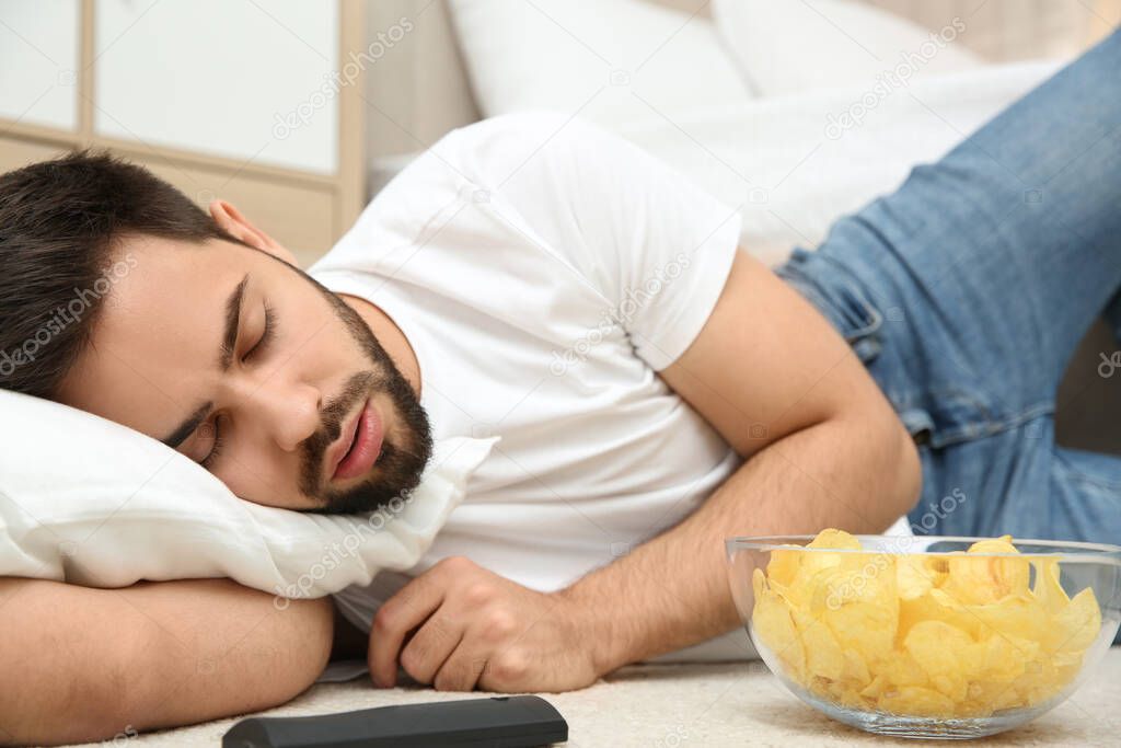 Lazy young man with bowl of chips and TV remote sleeping on floor at home