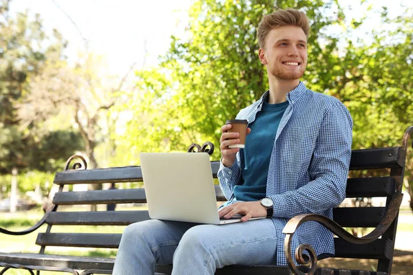 Young man with paper cup of coffee working on laptop in park
