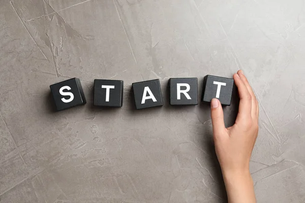 Woman making word START with black cubes on beige stone background, top view