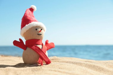 Kumsalda deniz kenarında Noel Baba şapkası ve eşarbıyla kumdan yapılmış bir kardan adam. Noel tatili