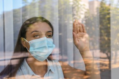 Camdan dışarı bakan koruyucu maskeli, stresli bir kadın. COVID-19 salgını sırasında kendini izole etme