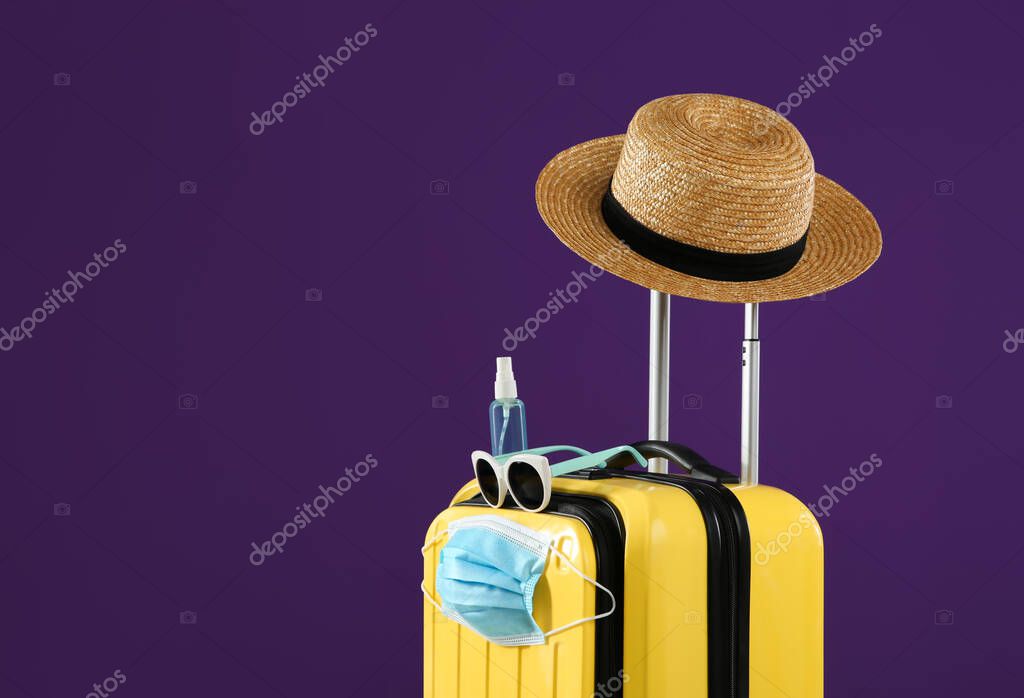 Stylish yellow suitcase with protective mask, sunglasses, hat and antiseptic spray on purple background, space for text. Travelling during coronavirus pandemic