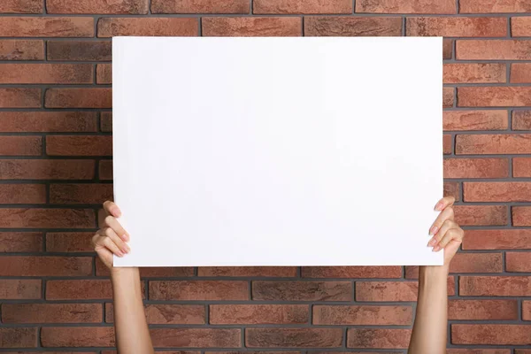 Woman holding white blank poster near red brick wall, closeup. Mockup for design