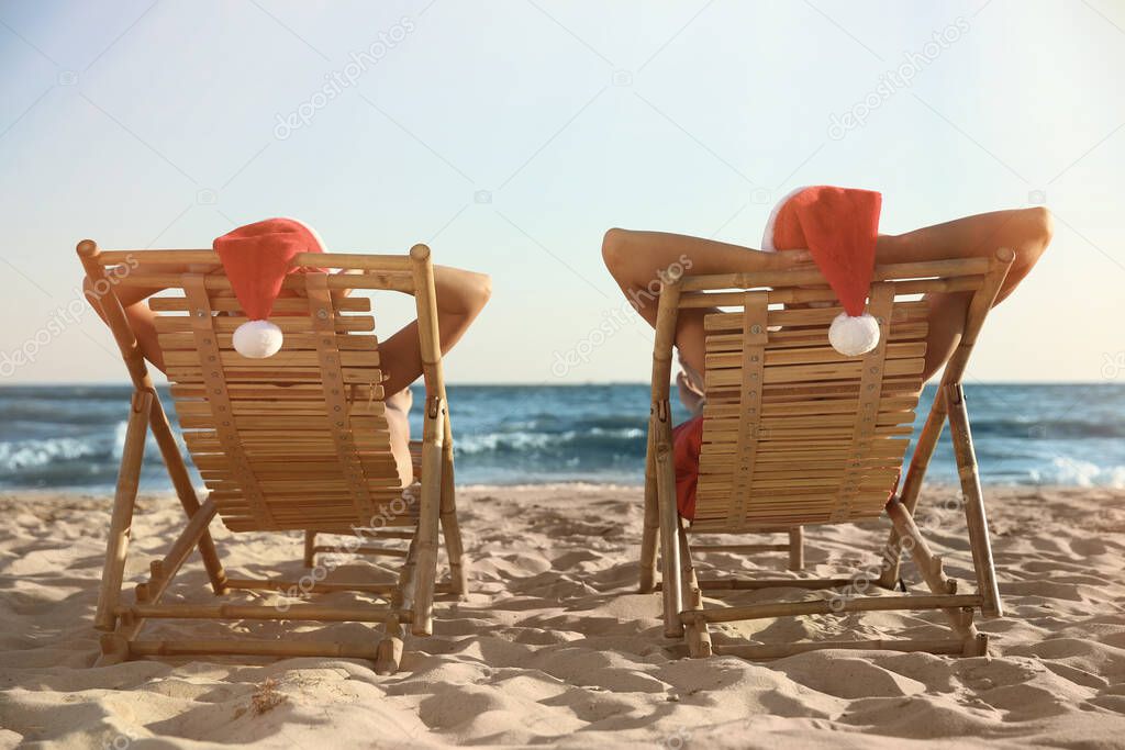 Lovely couple with Santa hats relaxing on deck chairs at beach, back view. Christmas vacation