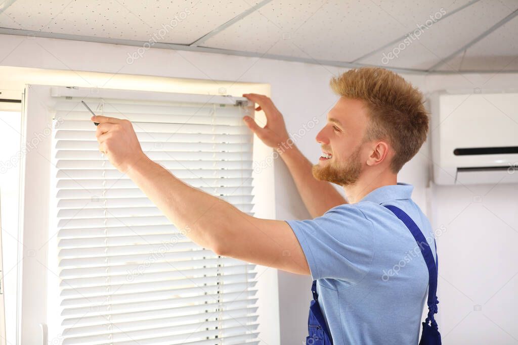 Handyman with screwdriver installing window blinds indoors