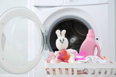 Çamaşır makinesinin yanındaki sepette bir şişe deterjan, oyuncak ve çocuk giysileri var.