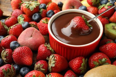 Fondue fork with strawberry in bowl of melted chocolate surrounded by other fruits on table clipart