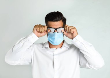 Hafif arka planda tıbbi maske takan bir adam sisli camları siliyor.