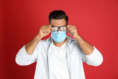 Kırmızı arka planda tıbbi maske takan bir adam sisli gözlük takıyor.