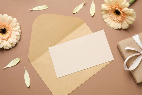 Beautiful herbera flowers, gift, envelope and blank card with space for text on brown background, flat lay