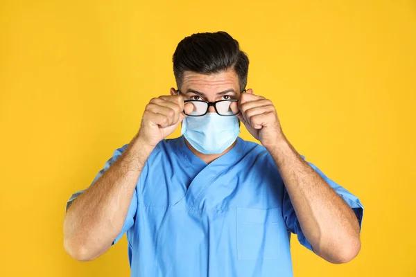 黄色の背景に医療用マスクを着用することによって引き起こされる霧の眼鏡を拭く医師 — ストック写真