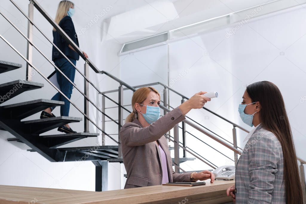 Woman in mask measuring temperature of employee with noncontact thermometer at office reception