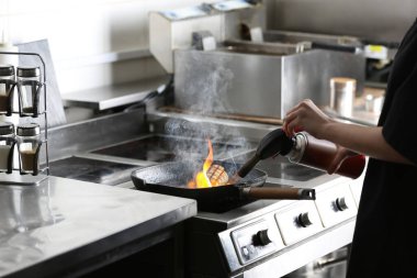 Kadın aşçı restoran mutfağında manuel gaz ocağıyla et pişiriyor.