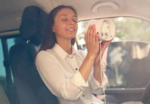 Young woman enjoying air flow from portable fan in car on hot summer day