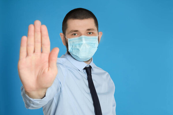 Man in protective mask showing stop gesture on light blue background, space for text. Prevent spreading of coronavirus