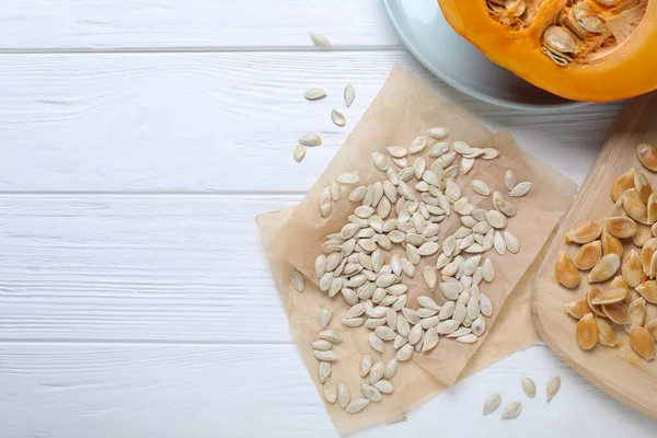 Raw seeds and fresh pumpkin on white wooden table, flat lay. Space for text