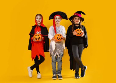 Balkabağı kovanlı şirin çocuklar sarı arka planda Cadılar Bayramı kostümleri giyiyorlar.