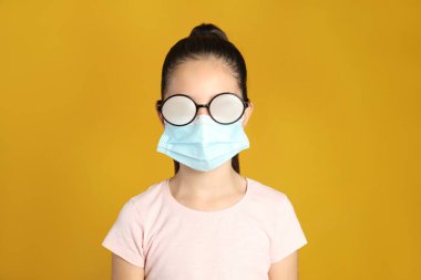Sarı arka planda tıbbi maske taktığı için sisli gözlüklü küçük bir kız. Koronavirüs salgını sırasında koruyucu tedbir