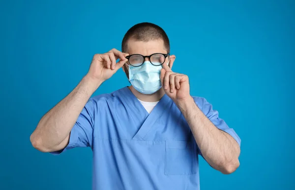 青色の背景に使い捨てマスクを着用することによって引き起こされる霧の眼鏡を拭く医師 コロナウイルスのパンデミック時の保護対策 — ストック写真