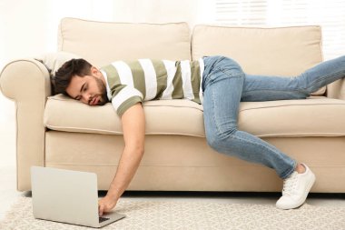 Lazy young man using laptop while lying on sofa at home clipart