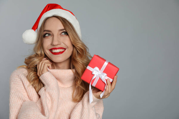 Happy young woman wearing Santa hat with Christmas gift on grey background. Space for text