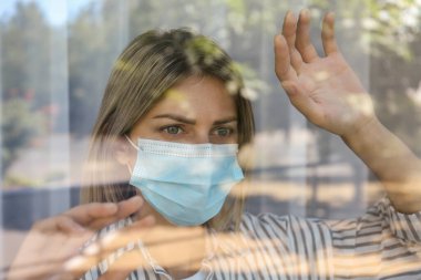 Camdan dışarı bakan koruyucu maskeli, stresli bir kadın. COVID-19 salgını sırasında kendini izole etme
