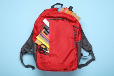 Açık mavi zemin üzerinde farklı kırtasiye malzemesi olan kırmızı sırt çantası, üst görünüm