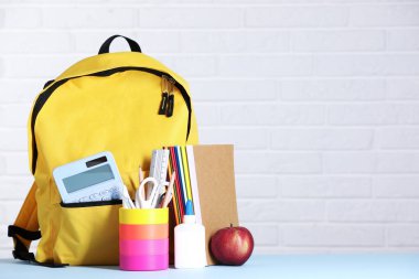 Sarı sırt çantası, farklı kırtasiye malzemeleri ve beyaz tuğla duvara karşı açık mavi masada elma. Metin için boşluk