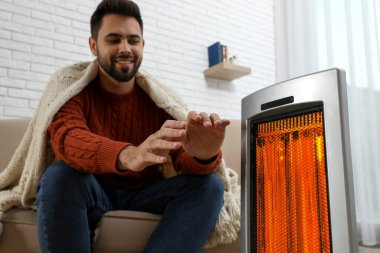 Adamın biri evde elektrikli ısıtıcının yanında ellerini ısıtıyor.