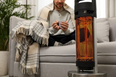 Evde elektrikli ısıtıcının yanında bir bardak sıcak içeceği olan adam seçici bir şekilde odaklanmış.