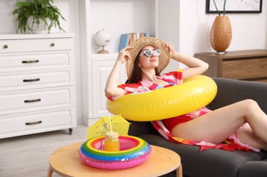 Güneş gözlüklü ve şapkalı şişme yüzüklü bir kadın evdeki kanepede eğleniyor. Denizde tatilin hayalini kuruyorum.