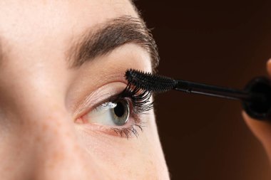 Genç bir kadın kahverengi arka planda kirpiklerine rimel sürüyor, yakın plan.