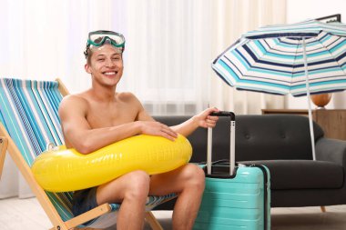 Dalış maskesi, bavulu ve evdeki katlanabilir sandalyede şişme yüzüğü olan mutlu bir adam. Denizde tatilin hayalini kuruyorum.