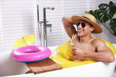 Güneş gözlüklü ve hasır şapkalı mutlu adam evdeki küvette şişme yüzüklü mason kavanozundan meyve suyu içiyor. Denizde tatilin hayalini kuruyorum.