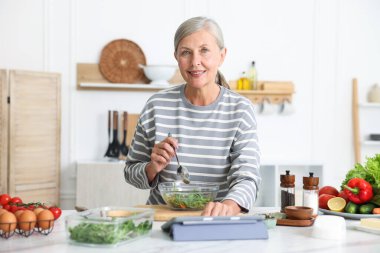 Mutfaktaki beyaz mermer masada yemek pişirirken tablet kullanan yaşlı kadın.