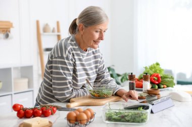 Mutfaktaki beyaz mermer masada yemek pişirirken tablet kullanan yaşlı kadın.