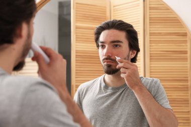 Evinde aynanın yanında yüzüne krem sürmekte zorlanan genç bir adam.