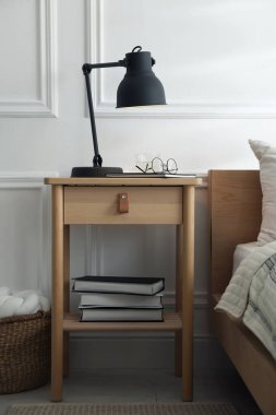 İçinde lamba, defter, gözlük, kitap ve yatağın yanında bir bardak su olan komodin.