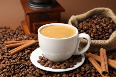 Kahverengi arka planda bir fincan sıcak kahve, fasulye, öğütücü ve tarçın, yakın plan.