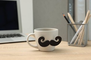 Babalar Günün kutlu olsun. Kağıt bıyıklı ve dizüstü bilgisayarlı bir fincan, ahşap masa, yakın plan.