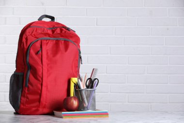 Kırmızı sırt çantası, farklı kırtasiye malzemeleri ve beyaz tuğla duvara karşı hafif mermer masa üzerinde elma. Metin için boşluk