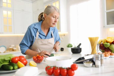Mutfaktaki beyaz mermer masada yemek pişirirken tablet kullanan yaşlı kadın.
