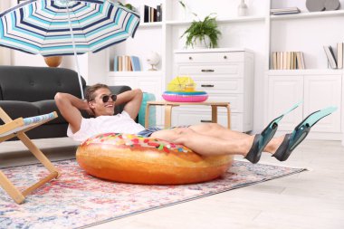 Güneş gözlüklü mutlu adam ve şişme yüzüklü paletler evde yerde eğleniyor. Denizde tatilin hayalini kuruyorum.