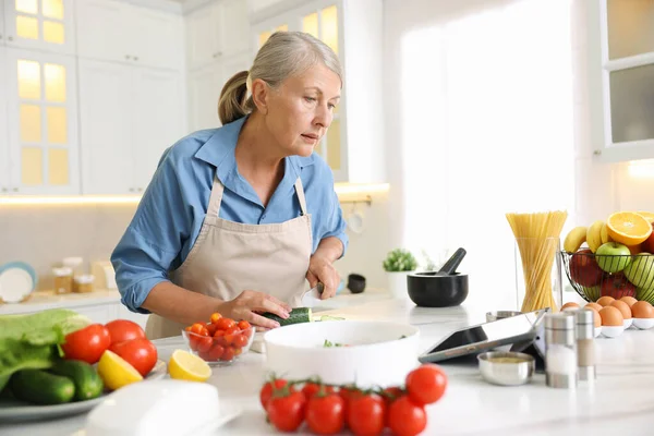 Mutfaktaki beyaz mermer masada yemek pişirirken tablet kullanan yaşlı kadın.